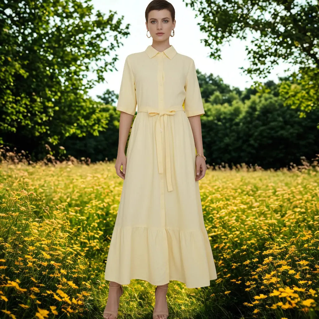 A woman stands in a sunlit field of yellow flowers wearing a pale yellow midi dress with short sleeves, a pointed collar, a front button placket, and a matching waist tie.