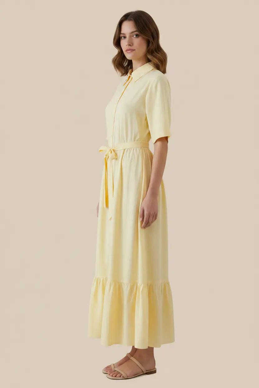 A woman modeling a pale yellow midi dress with short sleeves, a collar, a tie waist, and a ruffled hem, shown against a solid beige background.