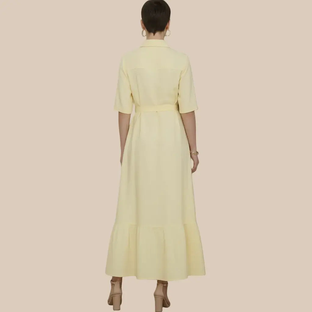 A woman viewed from the back wearing a pale yellow shirt dress with short sleeves, a collar, a matching waist tie, and a tiered hem, standing against a textured gray wall - Veyaro