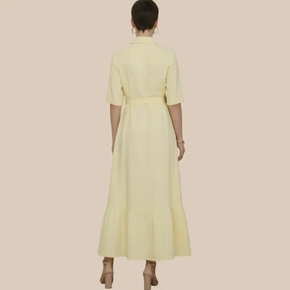 A woman viewed from the back wearing a pale yellow shirt dress with short sleeves, a collar, a matching waist tie, and a tiered hem, standing against a textured gray wall - Veyaro