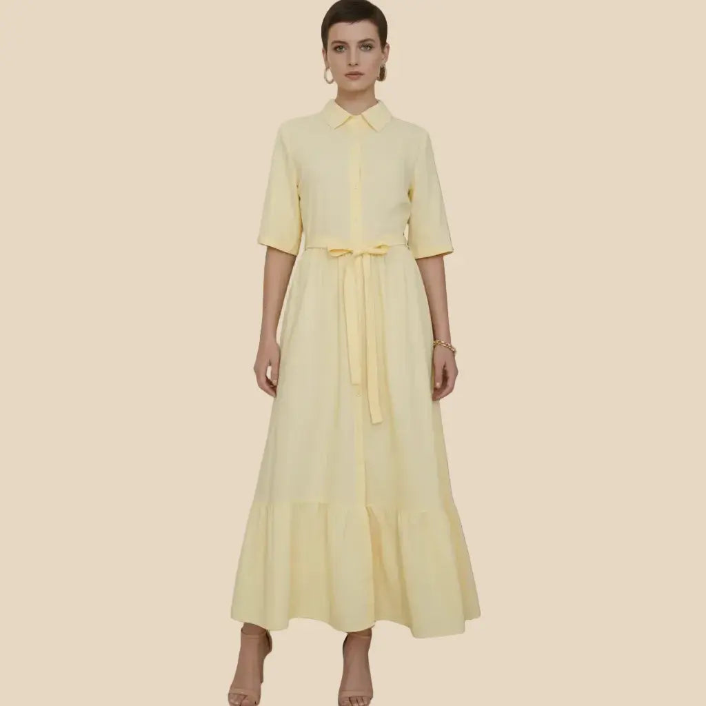 A woman modeling a pale yellow shirt dress with short sleeves, a pointed collar, a front button placket, and a matching waist tie, set against a solid beige background.