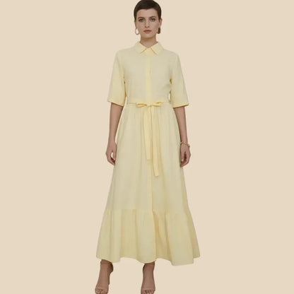 A woman modeling a pale yellow shirt dress with short sleeves, a pointed collar, a front button placket, and a matching waist tie, set against a solid beige background.
