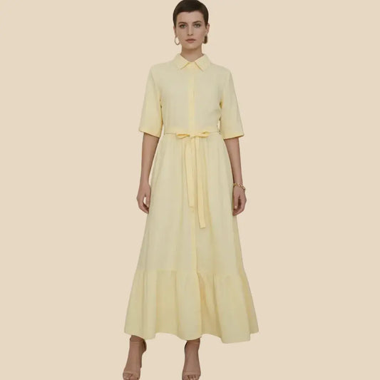 A woman modeling a pale yellow shirt dress with short sleeves, a pointed collar, a front button placket, and a matching waist tie, set against a solid beige background.