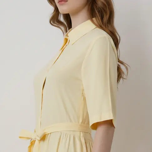A woman wearing a pale yellow short-sleeve collared shirt dress with a tie waist, shown in a side profile against a light neutral background.
