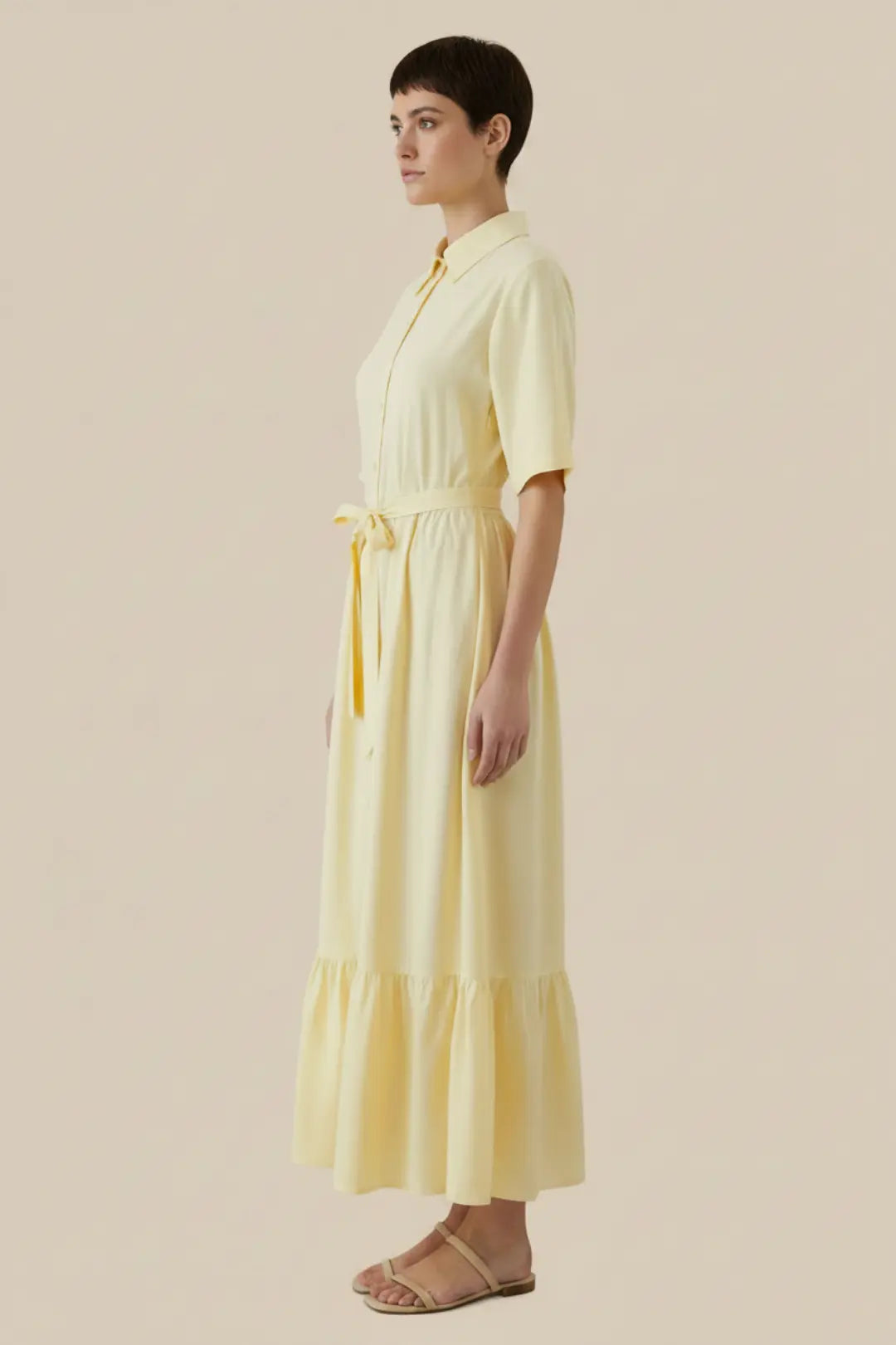 A woman modelling a pale yellow midi shirt dress with short sleeves, a button-front bodice, a matching waist tie, and a tiered skirt, shown in side profile against a plain beige studio background - Veyaro 