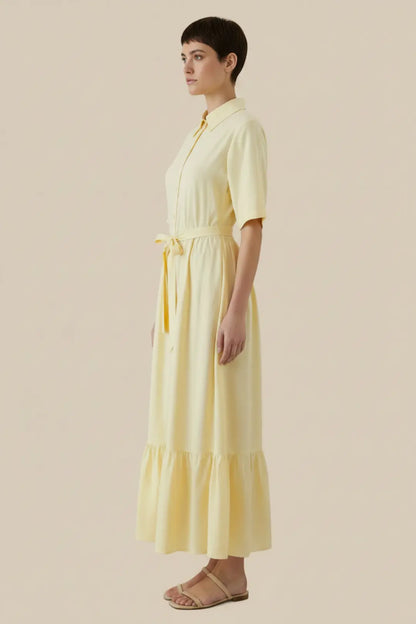 A woman modelling a pale yellow midi shirt dress with short sleeves, a button-front bodice, a matching waist tie, and a tiered skirt, shown in side profile against a plain beige studio background - Veyaro 