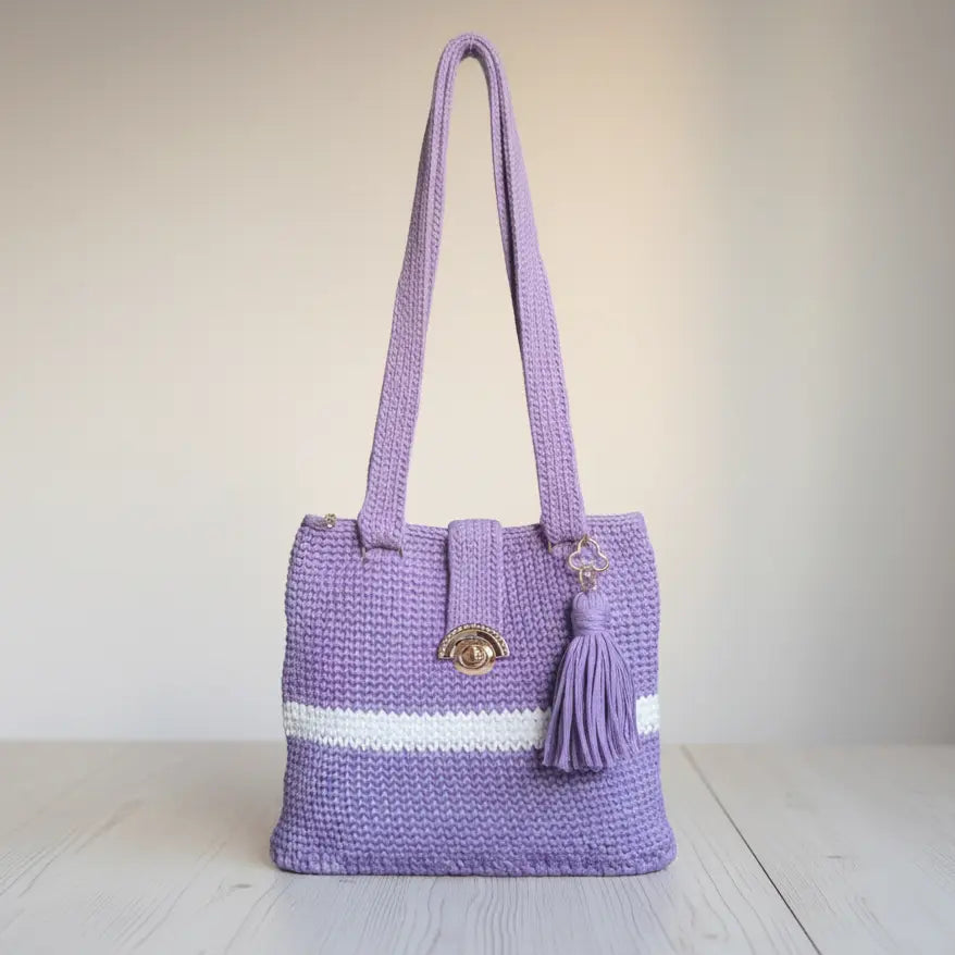 A lavender purple crocheted tote bag with a white stripe detail, a gold-tone oval clasp, and a matching purple tassel, set against a neutral beige background.