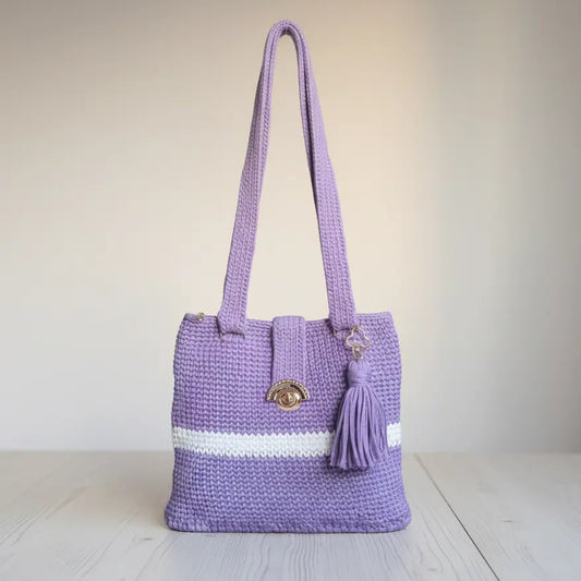 A lavender purple crocheted tote bag with a white stripe detail, a gold-tone oval clasp, and a matching purple tassel, set against a neutral beige background.
