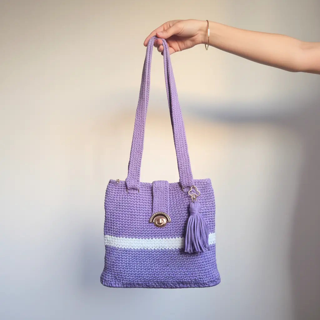A lavender purple crocheted tote bag with a white stripe and a tassel charm, held by a person's hand against a plain light grey background -Veyaro