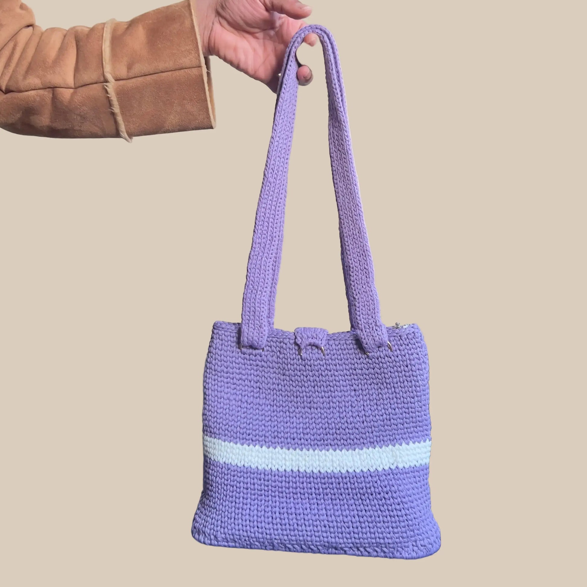 Purple knitted bag with a white stripe held by a person against a black leather couch.