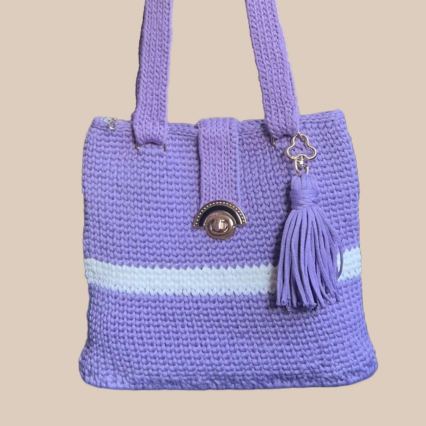 A lavender purple crocheted tote bag with a horizontal light blue stripe across the middle, featuring dual shoulder straps, a decorative metal clasp, and a matching purple tassel charm.