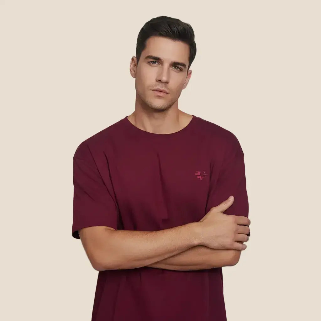A man wearing a short-sleeve crewneck t-shirt in deep burgundy with a small tonal embroidered logo on the left chest, posed against a neutral light gray background.
