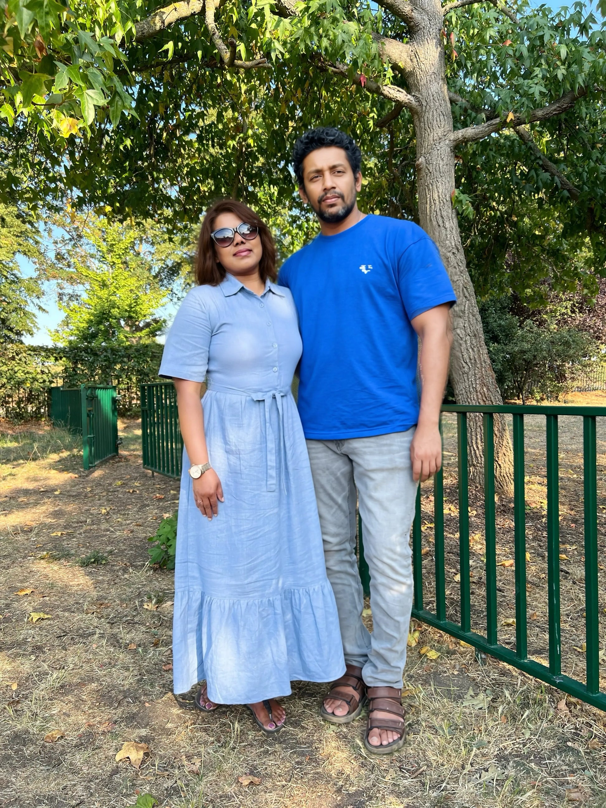 A man and a woman standing outdoors in a park-like setting with trees and a green fence. The woman wears a light blue short-sleeve midi dress, and the man wears a bright blue t-shirt, light gray jeans, and brown sandals.