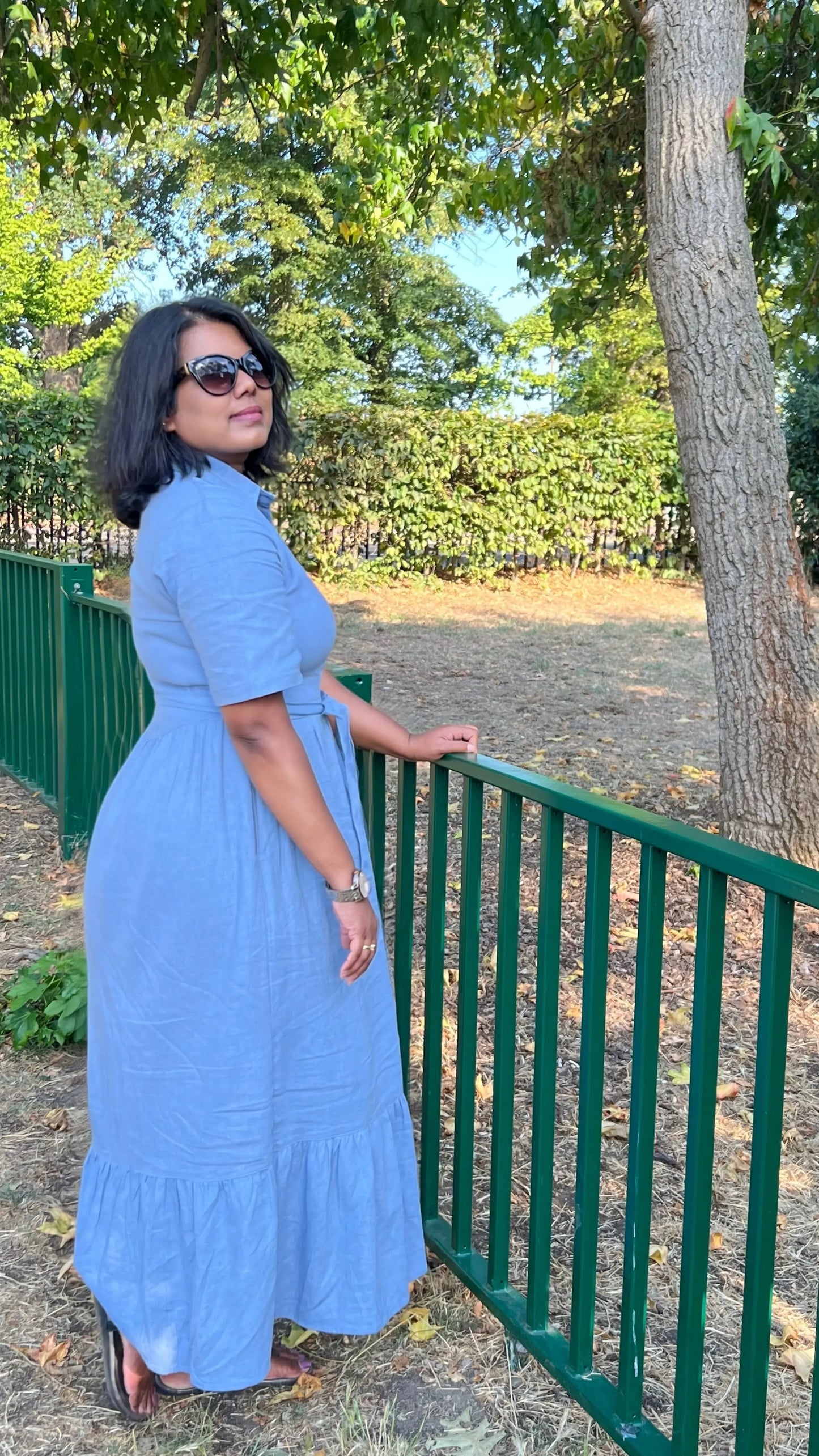 A woman wearing a light blue tiered midi dress and sunglasses stands outdoors next to a green metal fence, with trees and a grassy background.