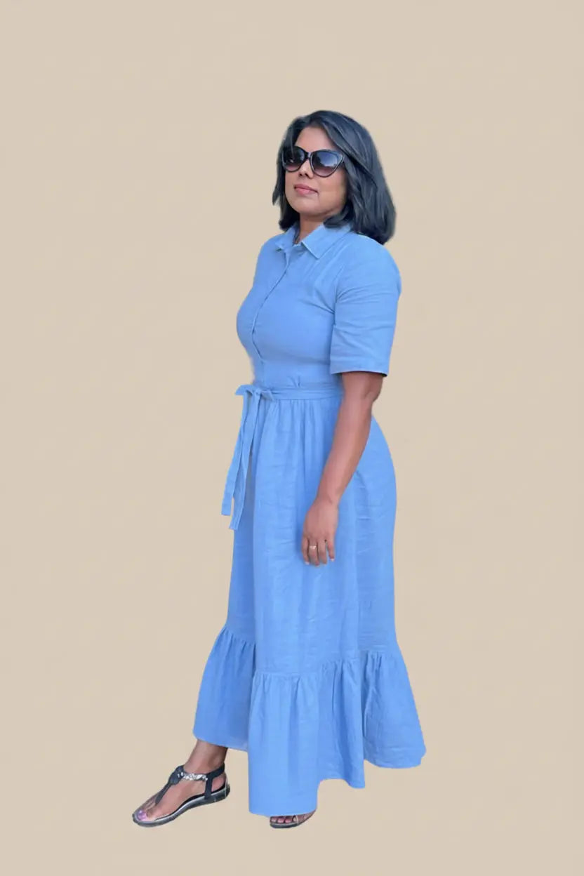 A woman wearing a light blue linen midi shirt dress with short sleeves, a button-up front, and a matching tie belt, standing outdoors next to a green metal fence.