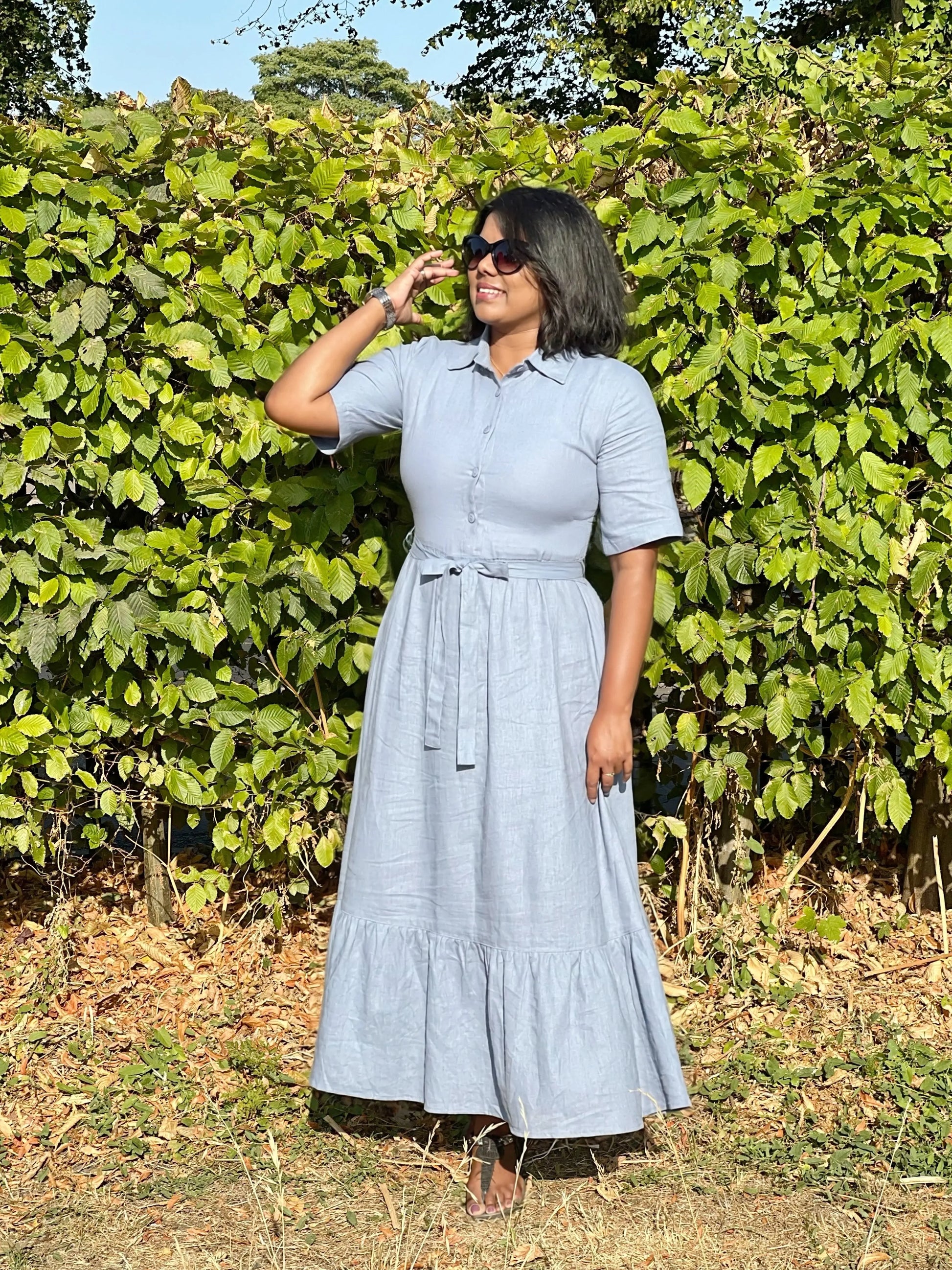 A woman wearing a light blue linen shirt dress with a tie waist and tiered skirt, standing outdoors in front of a green leafy hedge.