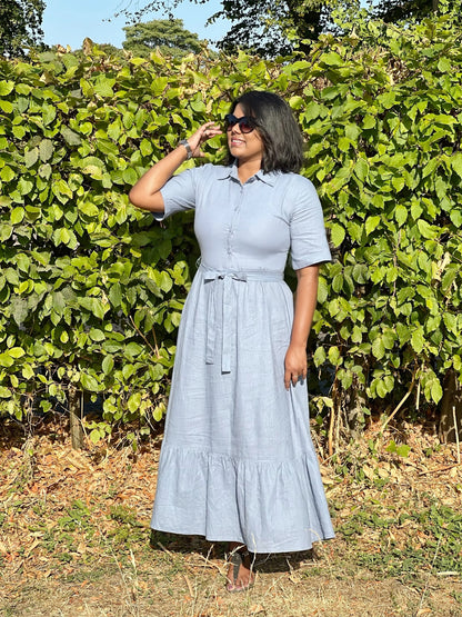 A woman wearing a light blue linen shirt dress with a tie waist and tiered skirt, standing outdoors in front of a green leafy hedge.