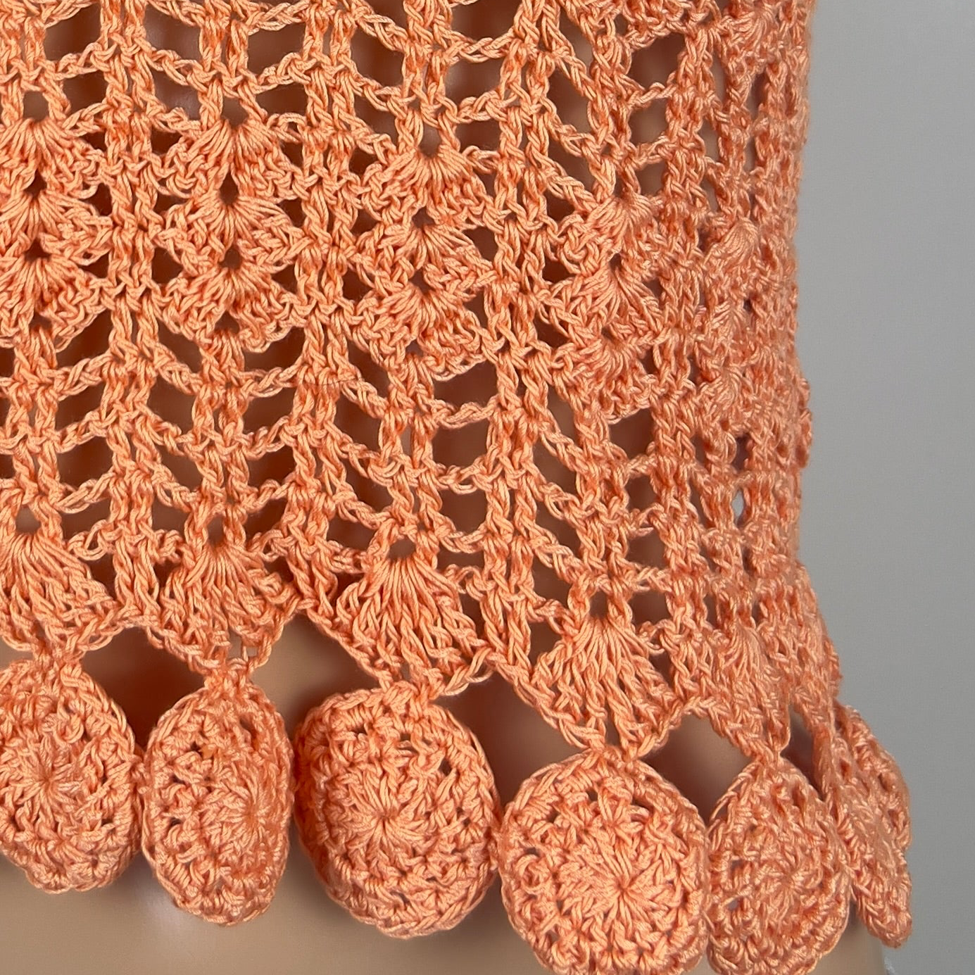 Close-up of a peach-colored crocheted fabric with circular patterns and tassels on a gray background.
