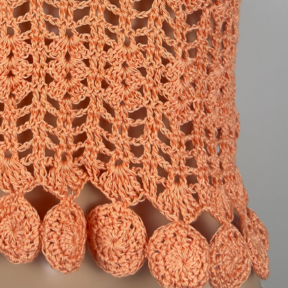 Close-up of a peach-colored crocheted fabric with circular patterns and tassels on a gray background.