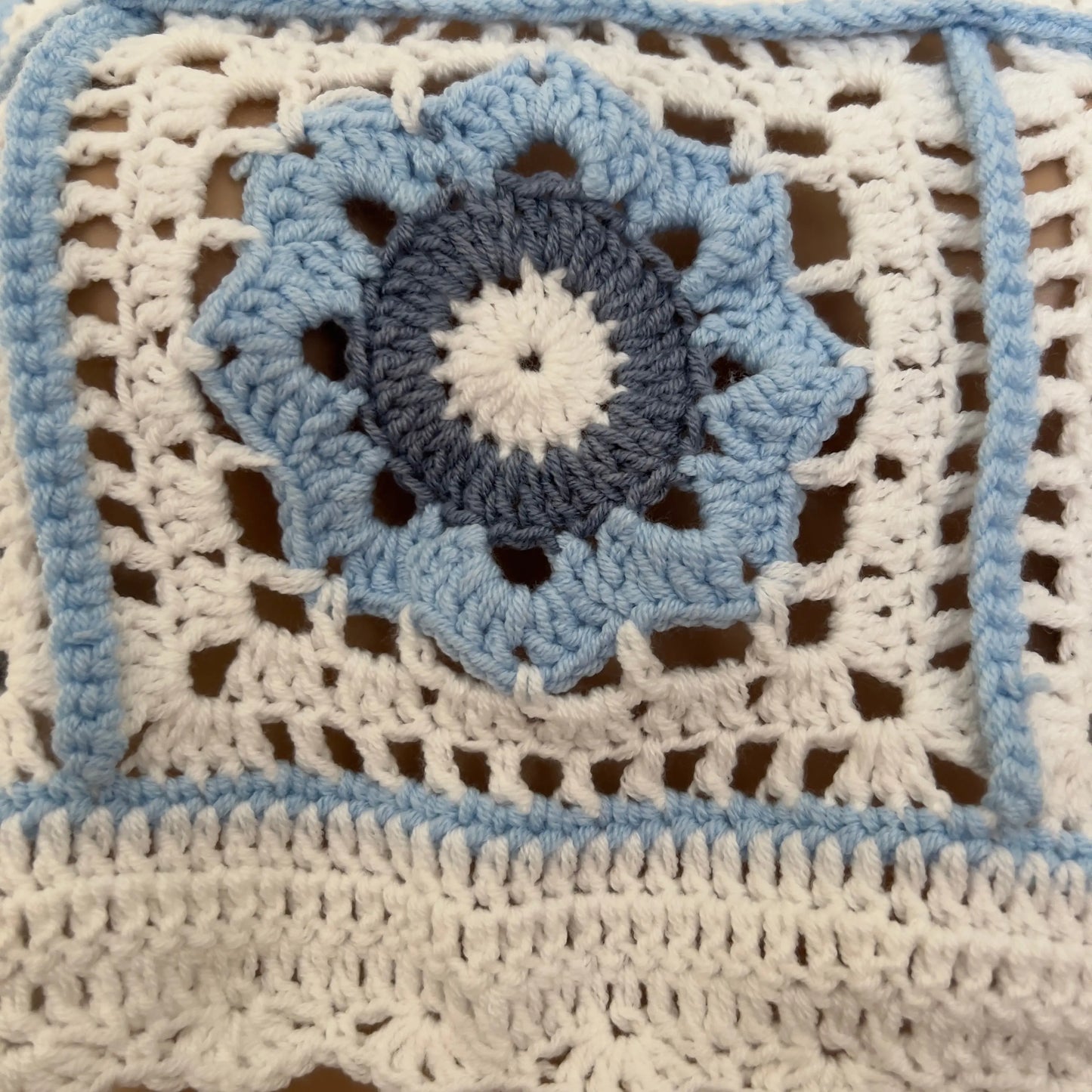 Close-up of a crocheted textile with a granny square design in cream, light blue, and dark blue, showing the detailed crochet pattern and openwork texture.